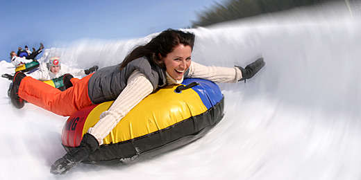 Eine Frau in Winterkleidung rutscht lächelnd auf einem bunten, aufblasbaren Schlauch einen verschneiten Hügel hinunter, während im Hintergrund andere Leute Schlauchboot fahren.