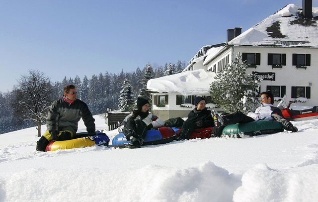 Vier Personen sitzen auf Snow Tubes in einer verschneiten Landschaft in der Nähe eines großen Gebäudes, mit schneebedeckten Bäumen und einem klaren Himmel im Hintergrund.