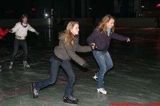 Drei junge Leute, die in einer Halle Schlittschuh laufen. Zwei Mädchen halten sich an den Händen und bewegen sich vorwärts, während ein anderes im Hintergrund Schlittschuh läuft.