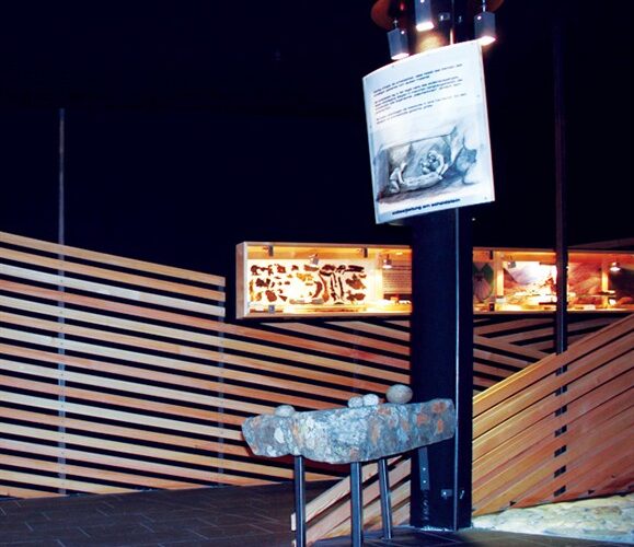 Eine Museumsausstellung zeigt ein Steinartefakt auf einem Ständer, eine Schautafel mit einem Bild und Text darüber und beleuchtete Vitrinen im Hintergrund.