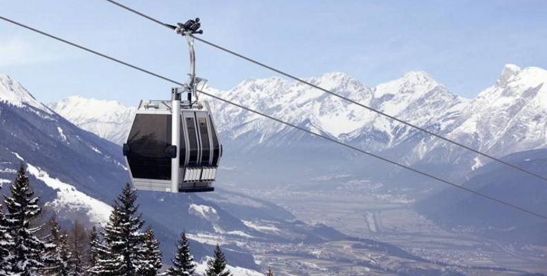 Eine Seilbahn fährt über eine verschneite Berglandschaft mit Kiefern und entfernten Tälern unter einem klaren Himmel.