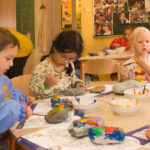 Drei kleine Kinder sitzen an einem Tisch in einem Klassenzimmer und bemalen große Felsen mit Pinseln und bunten Farben. Im Hintergrund sind andere Kinder und Kunstwerke zu sehen.
