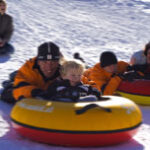 Drei Erwachsene und zwei Kinder fahren mit Snowtubes einen verschneiten Hügel hinunter, sie tragen Winterjacken und Helme. Eine weitere Person sitzt im Hintergrund.