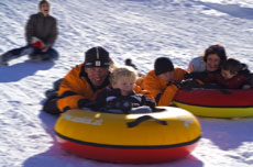 Drei Erwachsene und zwei Kinder fahren mit Snowtubes einen verschneiten Hügel hinunter, sie tragen Winterjacken und Helme. Eine weitere Person sitzt im Hintergrund.