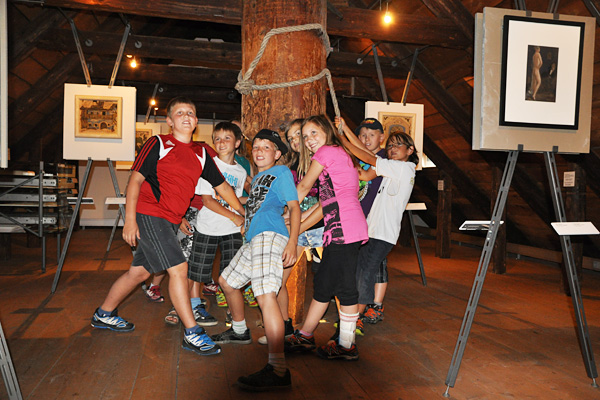 Eine Gruppe von Kindern steht in einer Kunstgalerie umher und lehnt sich an eine Holzsäule, während im Hintergrund gerahmte Kunstwerke auf Staffeleien ausgestellt sind.