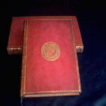 Zwei alte, in rotes Leder gebundene Bücher mit goldenen Verzierungen und einem geprägten Wappen auf dem vorderen Einband, die auf einer dunklen Fläche stehen.