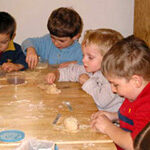 Vier kleine Kinder sitzen an einem Holztisch und formen mit ihren Händen und kleinen Werkzeugen Teig.