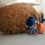 Drei Personen interagieren im Innenbereich mit einer großen, pelzigen, braunen, tunnelartigen Skulptur; zwei Kinder krabbeln heraus, während ein Erwachsener am Eingang lehnt.