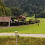 Ein Oldtimerzug fährt an Häusern vorbei und durch grüne Landschaften mit Bäumen und Hügeln im Hintergrund.