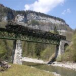 Ein schwarzer Dampfzug überquert eine grüne Metallbrücke über einen Fluss, mit felsigen Klippen und Bäumen im Hintergrund unter einem teilweise bewölkten Himmel.