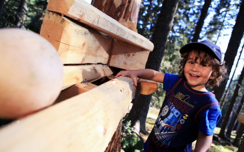 Ein kleines Kind in blauem Hemd und Mütze steht lächelnd an einem Holzgerüst im Freien, im Hintergrund sind Bäume zu sehen.