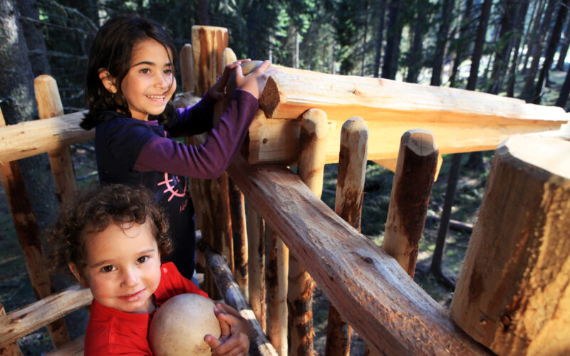 Zwei Kinder stehen in einer Holzkonstruktion in einem Wald; eines hält einen Ball und schaut in die Kamera, während das andere sich lächelnd am Geländer festhält.