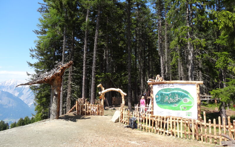 Ein hölzernes Eingangstor und ein Schild mit einer Landkarte stehen am Rande eines Waldes; zwei Personen stehen in der Nähe des Eingangs, im Hintergrund sind Berge zu sehen.