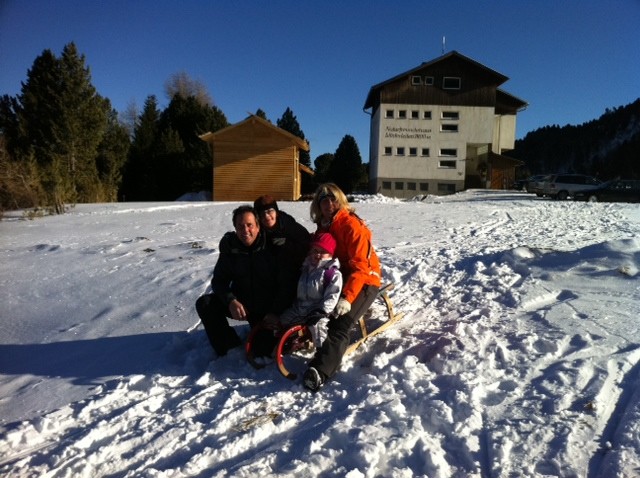 Vier Personen sitzen auf einem Holzschlitten im Schnee in der Nähe eines Gebäudes, mit Bäumen und einem klaren blauen Himmel im Hintergrund.