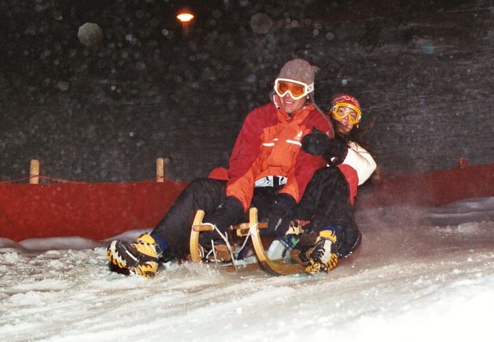 Zwei Personen in Winterkleidung und mit Schutzbrille fahren nachts auf einem Holzschlitten einen verschneiten Hang hinunter.