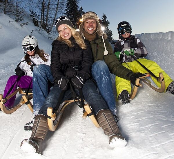 Eine vierköpfige Familie in Winterkleidung und mit Helmen fährt in der Abenddämmerung auf Holzschlitten einen verschneiten Hügel hinunter.