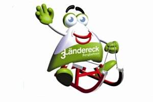 Cartoon-Maskottchen mit grün-weißem, dreieckigem Körper und der Aufschrift "3-Ländereck Bergbahnen", das lächelnd und winkend auf einem roten Schlitten sitzt.