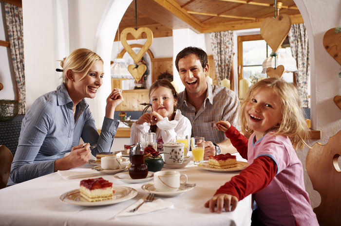 Eine vierköpfige Familie sitzt an einem Tisch in einem gemütlichen Restaurant, lächelt und genießt gemeinsam Kuchen und Getränke. Die Umgebung ist mit Holzdekor und herzförmigen Ornamenten geschmückt.