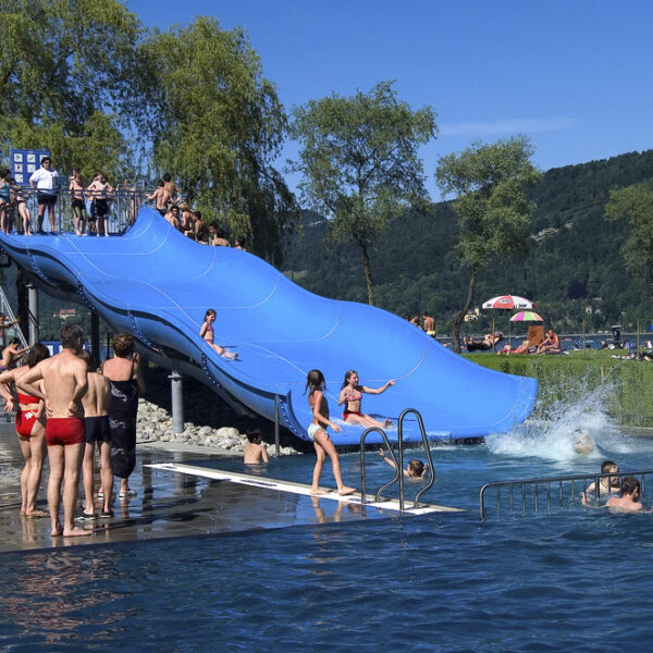 Menschen genießen einen sonnigen Tag in einem überfüllten Freibad mit einer großen blauen Dreifachrutsche, umgeben von Bäumen und Hügeln.
