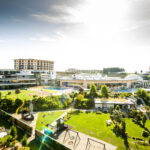 Ein großer Ferienkomplex mit mehreren Gebäuden, Außenpools, Gärten, Rasenflächen und Erholungsgebieten unter einem hellen, sonnigen Himmel.