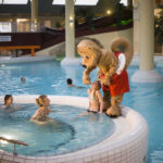 Eine Person in einem Eichhörnchen-Maskottchen-Kostüm steht am Rande eines Hallenbades und unterhält sich mit einem Kind, während andere in der Nähe schwimmen.