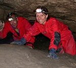 Zwei Personen in roten Overalls und mit Scheinwerfern kriechen durch einen engen Höhlengang und lächeln in die Kamera.