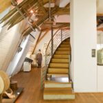 Innenraum eines modernen Museums mit Holzböden, sichtbaren Balken, Vitrinen, Wandausstellungen und einer Treppe, die in ein höheres Stockwerk führt.