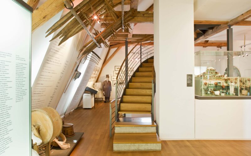 Innenraum eines modernen Museums mit Holzböden, sichtbaren Balken, Vitrinen, Wandausstellungen und einer Treppe, die in ein höheres Stockwerk führt.