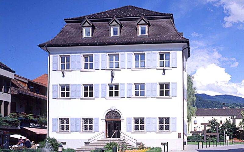 Ein weißes dreistöckiges Gebäude mit blauen Fensterläden, einem dunklen Ziegeldach und symmetrischen Fenstern, umgeben von Blumen und Grünpflanzen unter einem teilweise bewölkten Himmel.