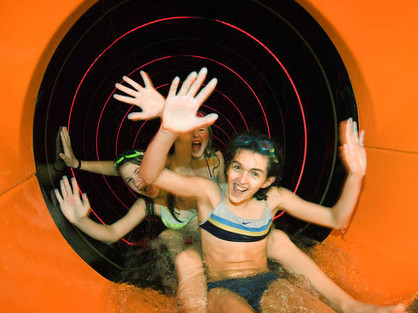 Drei Kinder in Badeanzügen verlassen eine orangefarbene Wasserrutsche, lächeln und winken aufgeregt mit den Händen.