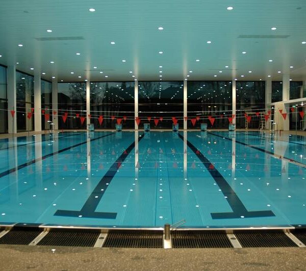 Ein Hallenbad mit Bahnmarkierungen und roten Fahnen, die über dem Wasser hängen; große Fenster säumen die Wände und die Deckenbeleuchtung spiegelt sich auf der Wasseroberfläche.