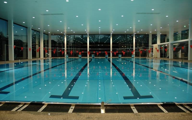 Ein Hallenbad mit Bahnmarkierungen und roten Fahnen, die über dem Wasser hängen; große Fenster säumen die Wände und die Deckenbeleuchtung spiegelt sich auf der Wasseroberfläche.