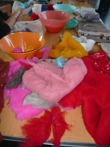 Bunte Wollreste und Filzstücke werden auf einem Tisch mit Schüsseln, Tassen und Bastelmaterial in einer Werkstattumgebung ausgebreitet.