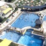 Luftaufnahme eines öffentlichen Freibadkomplexes mit Wasserrutschen, mehreren Becken, Sonnenliegen und grünen Rasenflächen.