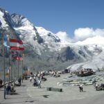Die Touristen versammeln sich auf einer Aussichtsplattform mit mehreren Fahnen und blicken auf schneebedeckte Berge und einen Gletscher unter einem klaren blauen Himmel.