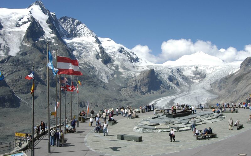 Die Touristen versammeln sich auf einer Aussichtsplattform mit mehreren Fahnen und blicken auf schneebedeckte Berge und einen Gletscher unter einem klaren blauen Himmel.