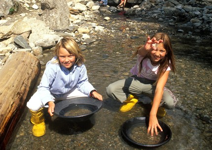 Zwei Kinder knien in einem flachen, felsigen Bach und halten jeweils eine schwarze Pfanne ins Wasser, wahrscheinlich um nach Gold zu wühlen.