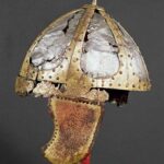 Ein korrodierter antiker Helm mit kuppelförmigem Metallrahmen, Ziernieten und beschädigtem Wangenschutz, ausgestellt auf einem roten Ständer vor einem schlichten Hintergrund.