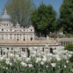 Eine detailgetreue Miniaturnachbildung des Petersdoms steht umgeben von Bäumen und blühenden weißen Tulpen in einer Parklandschaft.