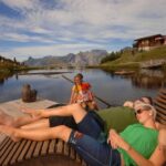 Zwei Erwachsene entspannen sich auf hölzernen Liegestühlen an einem See, während ein Kind in der Nähe sitzt. Im Hintergrund sind Berge, ein hölzerner Steg und ein Gebäude bei teilweise bewölktem Himmel zu sehen.