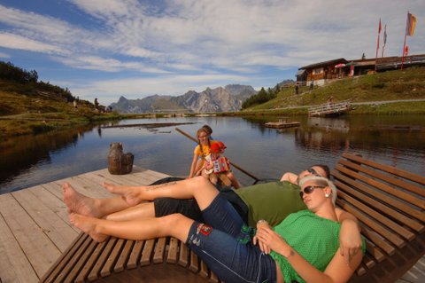 Zwei Erwachsene entspannen sich auf hölzernen Liegestühlen an einem See, während ein Kind in der Nähe sitzt. Im Hintergrund sind Berge, ein hölzerner Steg und ein Gebäude bei teilweise bewölktem Himmel zu sehen.