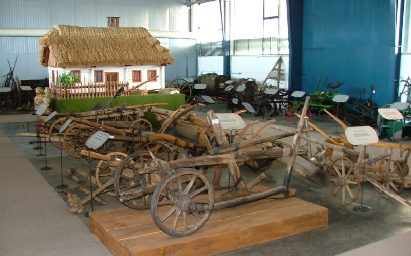 In den Innenräumen sind auf Podesten mehrere alte hölzerne landwirtschaftliche Geräte und Pflüge ausgestellt, mit einem Modell eines strohgedeckten Bauernhauses im Hintergrund.