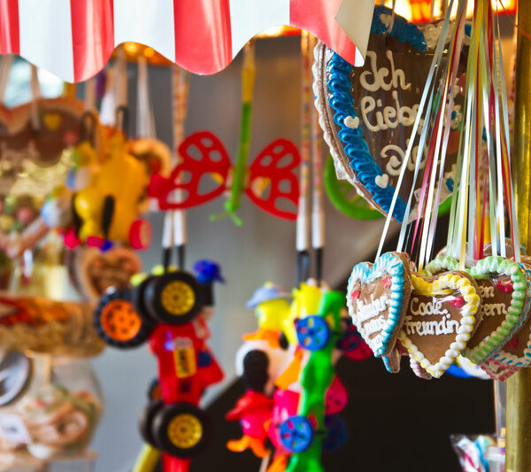 An einem bunten Marktstand werden unter einem gestreiften Vordach verzierte Lebkuchenherzen mit deutschen Texten, Spielzeugautos und Scherzbrillen angeboten.
