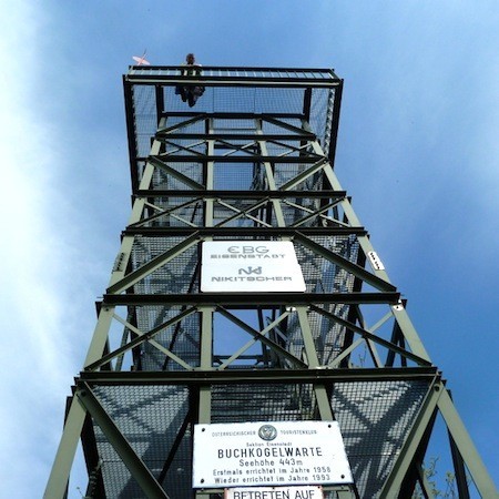 Ein metallener Aussichtsturm mit Informationsschildern an seiner Struktur wird von unten gegen einen blauen Himmel fotografiert. Eine Person steht auf der obersten Plattform.