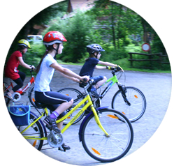 Drei Kinder mit Helmen fahren mit dem Fahrrad auf einem gepflasterten Weg in einem bewaldeten Außenbereich.