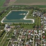 Luftaufnahme eines rechteckigen Sees mit einer kleinen Insel, umgeben von grünen Feldern und angrenzend an einen Campingplatz mit Reihen von Wohnwagen und Hütten.