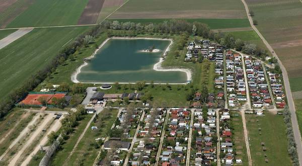 Luftaufnahme eines rechteckigen Sees mit einer kleinen Insel, umgeben von grünen Feldern und angrenzend an einen Campingplatz mit Reihen von Wohnwagen und Hütten.