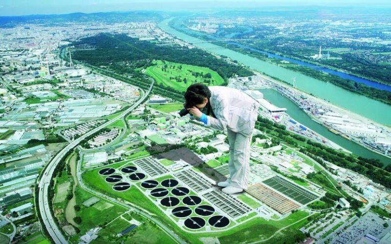 Ein digital bearbeitetes Bild einer Person in weißer Kleidung, die riesengroß erscheint, über einer Luftaufnahme einer Stadt steht und die Landschaft darunter fotografiert.