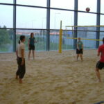 Sechs Personen spielen Volleyball auf einem überdachten Sandplatz mit großen Fenstern im Hintergrund.