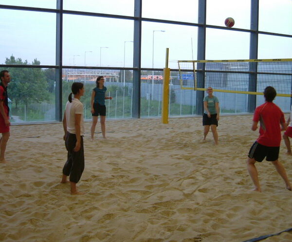 Sechs Personen spielen Volleyball auf einem überdachten Sandplatz mit großen Fenstern im Hintergrund.
